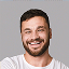 Uśmiechnięty mężczyzna z ciemnymi, krótkimi włosami i zarostem, ubrany w biały T-shirt, pozuje do zdjęcia na jednolitym szarym tle.