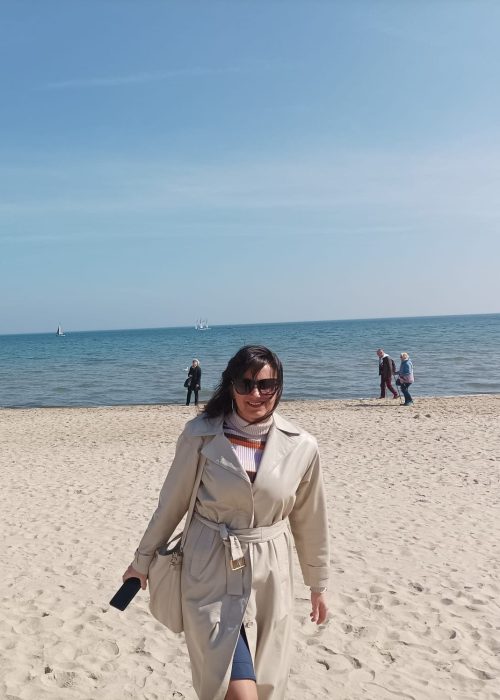 Kobieta w beżowym trenczu i okularach przeciwsłonecznych idzie po piaszczystej plaży w słoneczny dzień, trzymając w dłoni telefon. Za nią rozciąga się morze, a w oddali widać kilka niewyraźnych postaci i żaglówki.